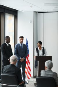A diverse group of professionals in a formal meeting, with speakers at a podium.