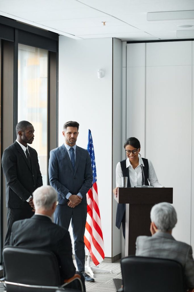 A diverse group of professionals in a formal meeting, with speakers at a podium.